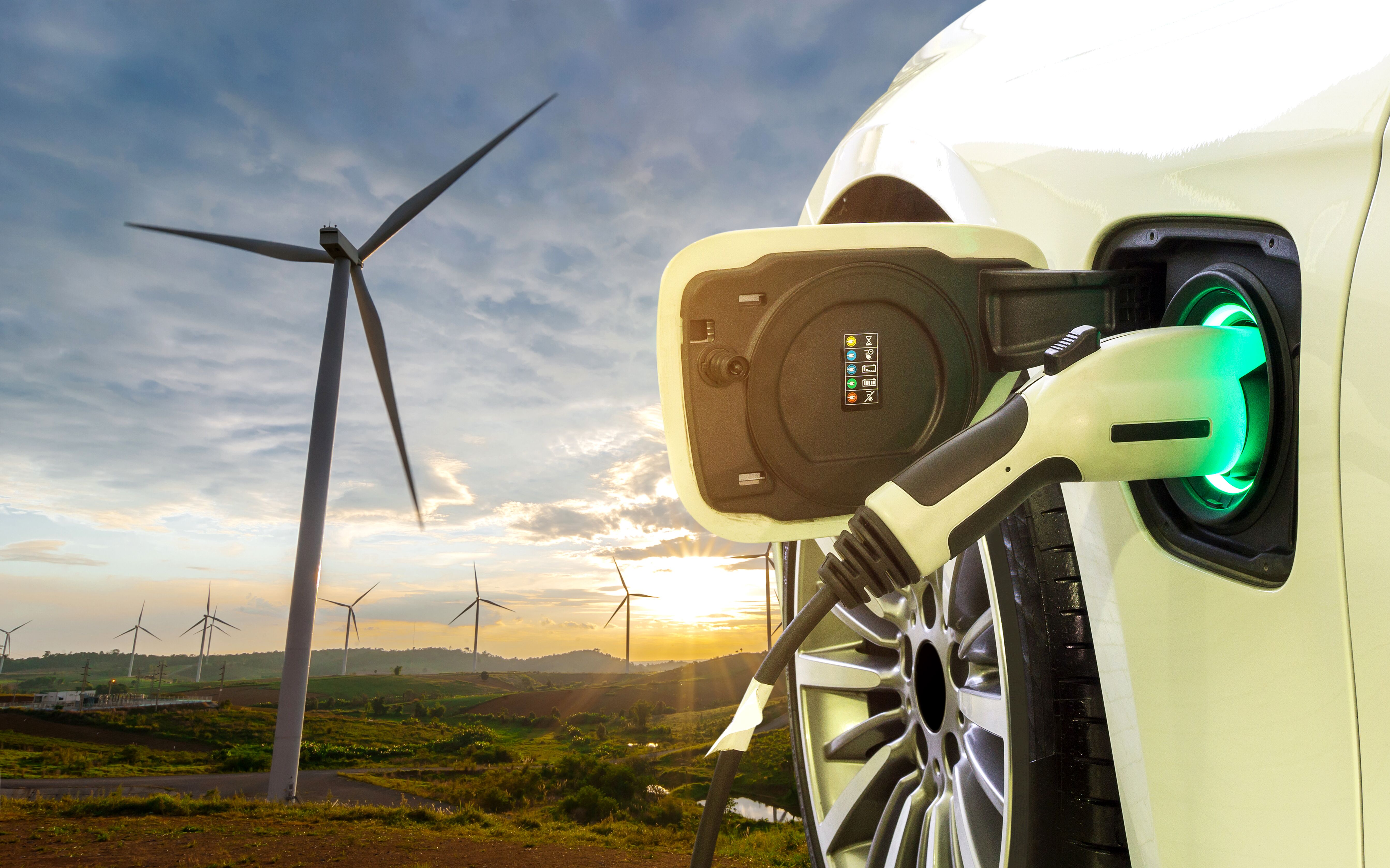Image of a wind farm with the sun setting behind it with an electric vehicle in the foreground with its electrical port open and a charging cord plugged in